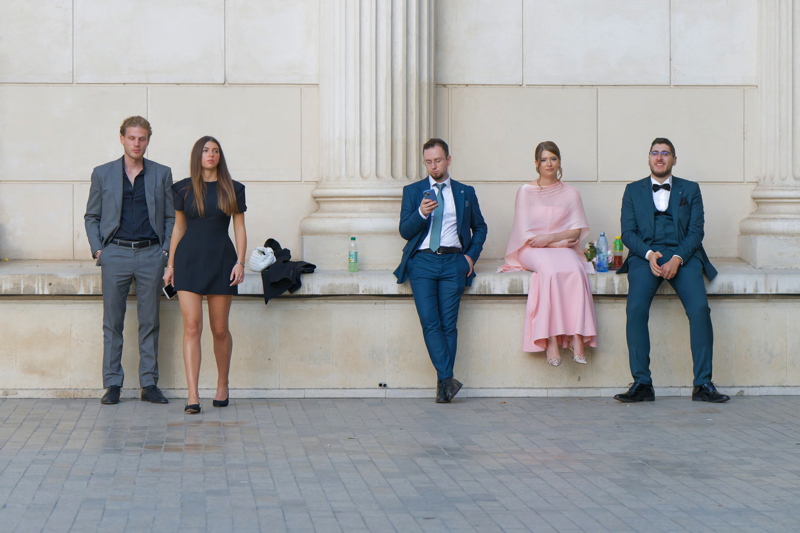 16 - Five young people dressed in formal attire including suits and dresse, sitting and standing against a stone wall with classical columns